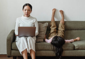 Woman using a laptop with her child sitting next to her, but the child is upside down on the couch.