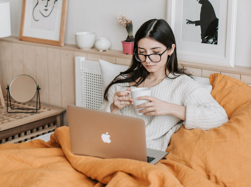 Woman using her apple laptop in her bed while holding a cup of coffee.
